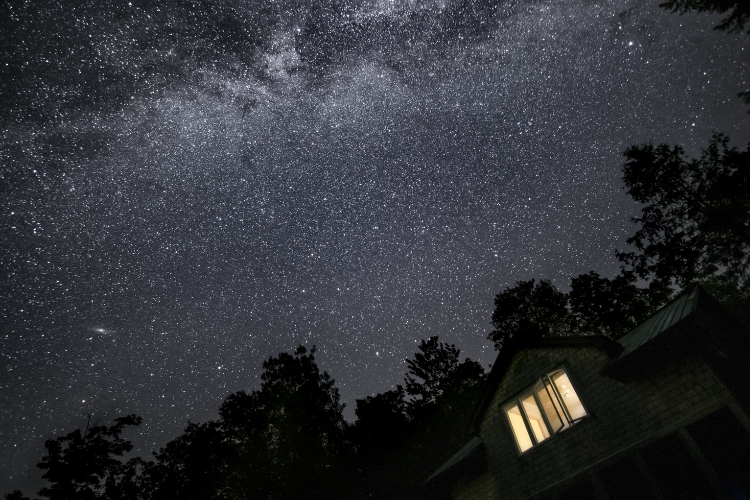 The milky way over some trees and the top of a wooden cabin with a lit window